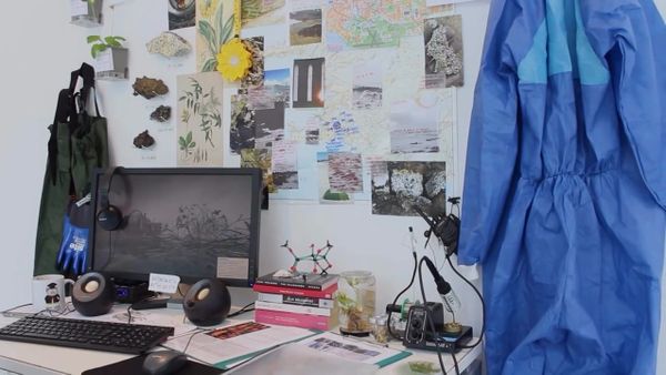 An installation desk crowded with specimens, botanical clippings, a monitor and blue lab coveralls