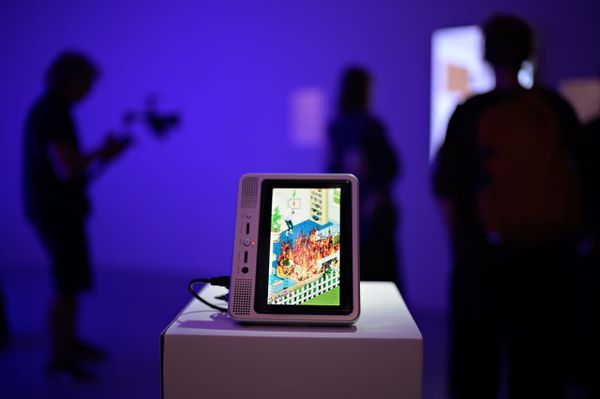 A portable DVD player on a white plinth shows a Sims house surrounded by flames, with visitors in silhouette behind it