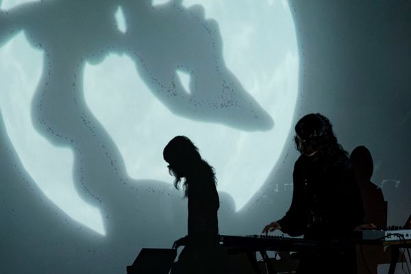 Two performers in black at a table of synthesizers, silhouetted against a projected moon crossed by a large shadow.