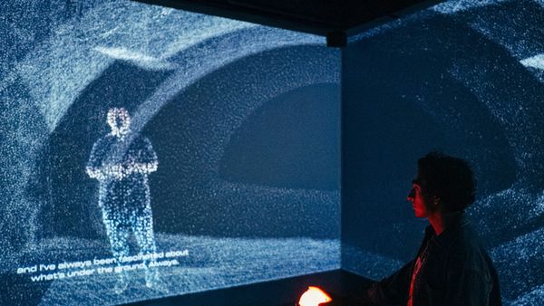 A visitor rests a hand on a glowing red orb in a dark room where a tunnel and a human figure are projected as clouds of blue-white points.