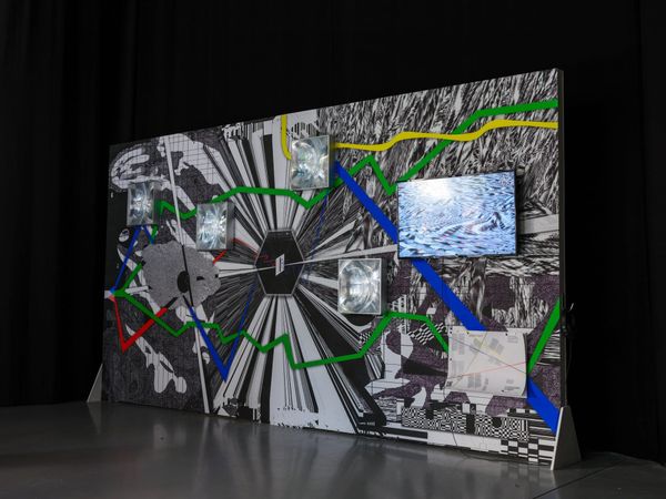A freestanding wall printed with black-and-white patterns and crossed by red, green, blue and yellow lines, with a video screen and four square glass panels mounted on it.