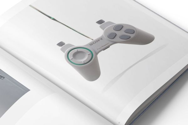 An open book page shows a photograph of a grey prototype PlayStation controller with a green ring around its directional pad.