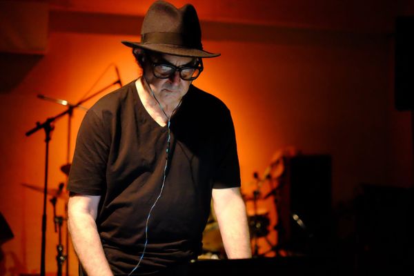 Carl Stone, wearing a hat and glasses, performs in front of an orange-lit wall.