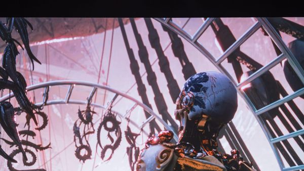 A projected CGI scene shows a figure with a blue-patterned porcelain head, seen from behind, inside an iron and glass structure.