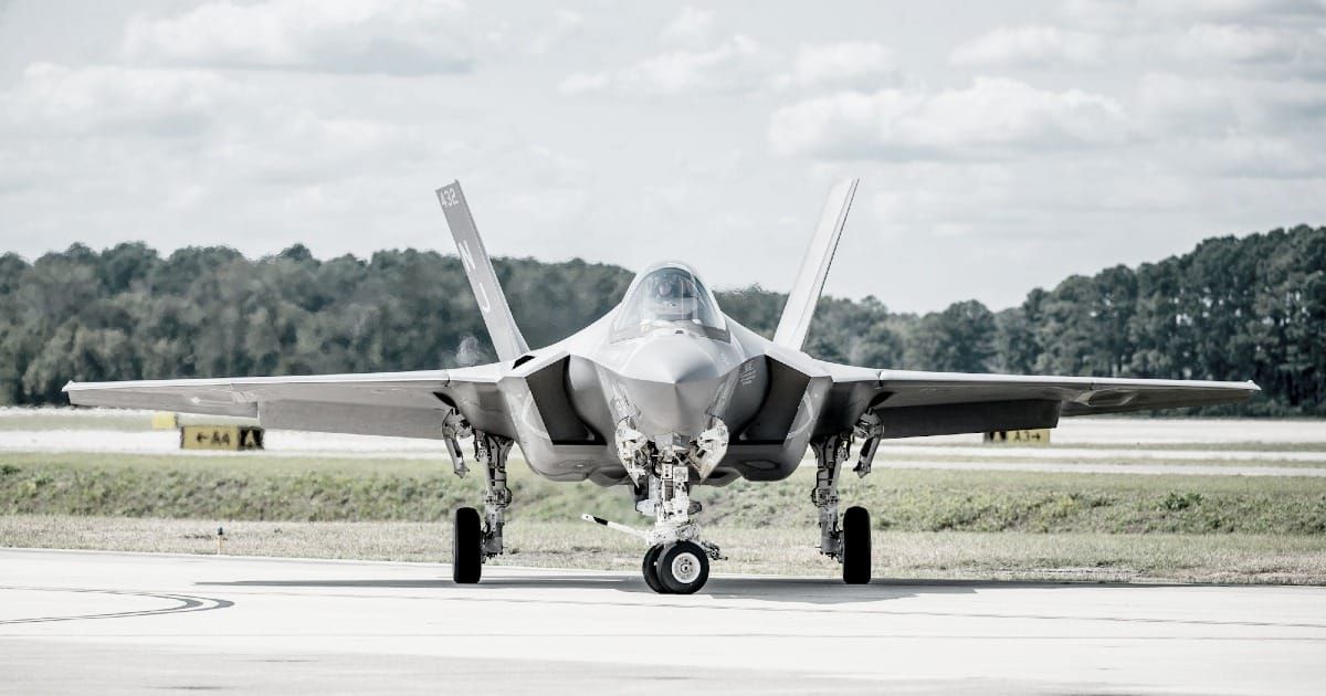 Frontal view of an F-35 Lightning II stealth fighter jet. It is positioned on an airfield or taxiway during the day, under a slightly overcast sky with soft, diffused lighting.