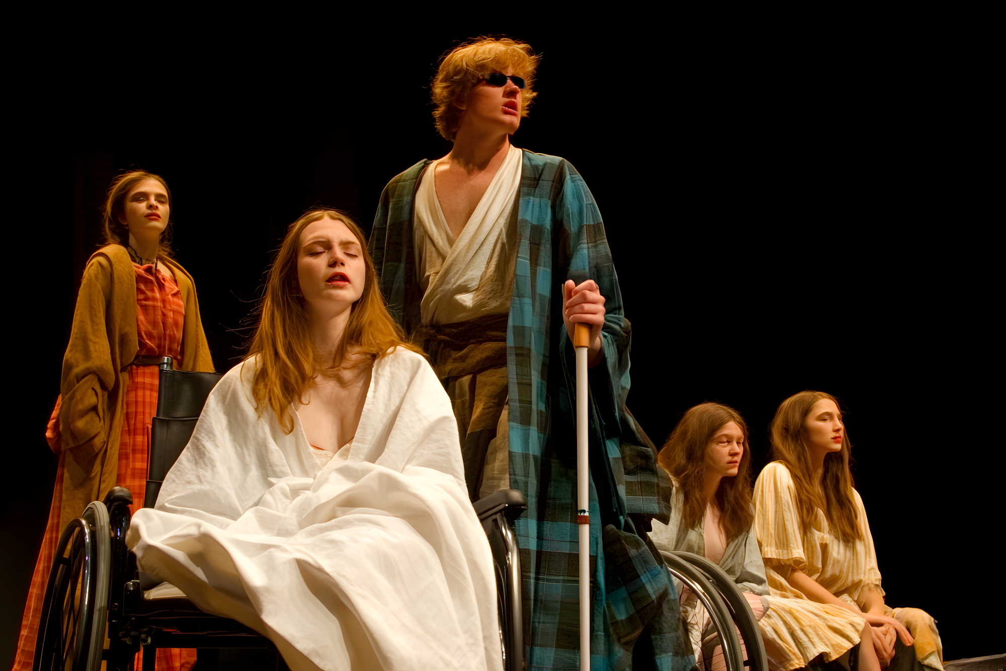 Sur une scène de théâtre plongée dans le noir, cinq personnes sont disposées dans une composition dramatique. Au centre, une personne en fauteuil roulant porte une robe blanche ample, la tête légèrement renversée en arrière. À ses côtés, une personne debout s'appuie sur une canne blanche et porte une robe de chambre à carreaux turquoise. Trois autres personnes complètent la scène, vêtues de costumes d'époque dans des tons jaune et beige. L'éclairage théâtral crée une ambiance rappelant une peinture classique, avec des contrastes marqués entre ombre et lumière.