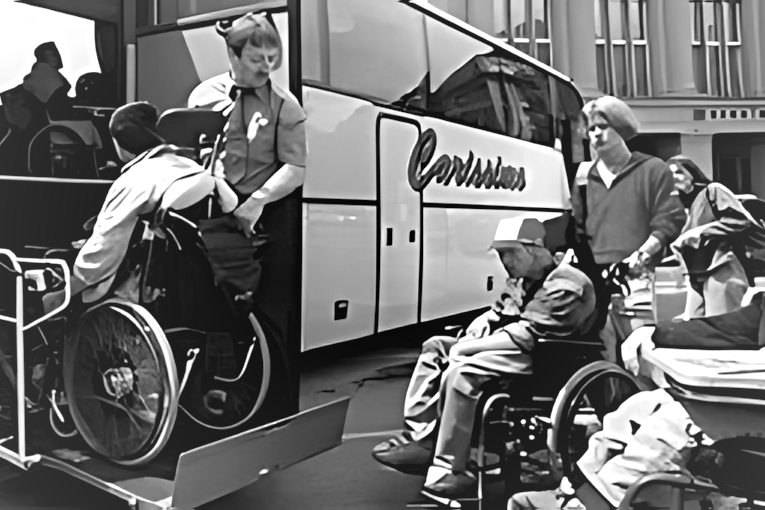 Photographie en noir et blanc de 2002, de mauvaise qualité, montrant l'embarquement dans un autocar accessible. Deux personnes en fauteuil roulant utilisent un élévateur hydraulique : l'une est sur la plateforme en cours de montée, l'autre attend au sol. Le chauffeur en uniforme et du personnel d'accompagnement supervisent l'opération.