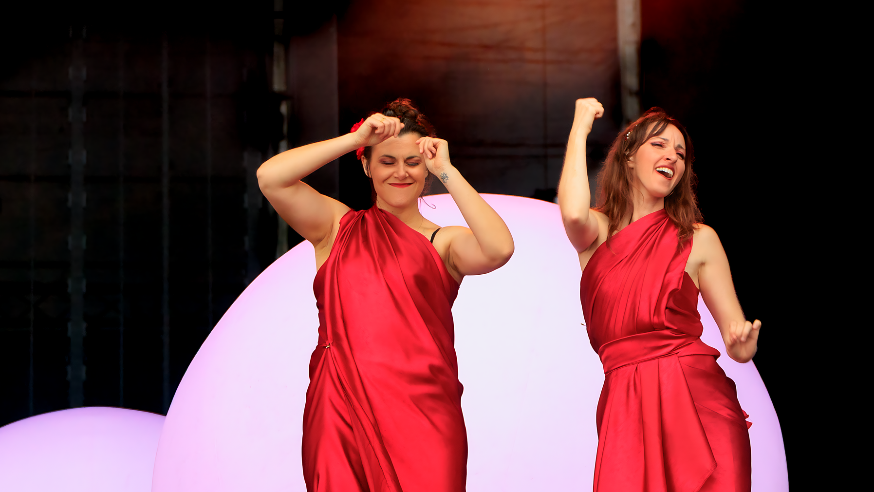Deux femmes en robes rouges drapées performent sur scène devant un décor rose et violet. Perinne Diot (à gauche) pose ses mains sur sa tête dans un geste théâtral, tandis qu'Aurélie Nahon (à droite) lève le poing en l'air, toutes deux souriant lors de leur interprétation en langue des signes des chansons de Philippe Katerine aux Vieilles Charrues.