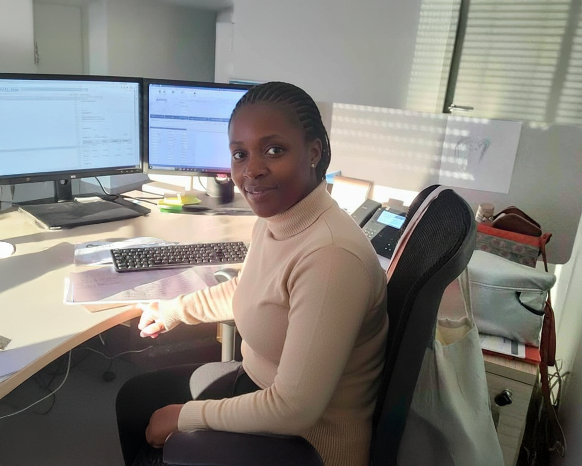 Une femme, assise à un poste de travail de bureau devant deux écrans d'ordinateur allumés et un clavier, regarde en direction de l'objectif.