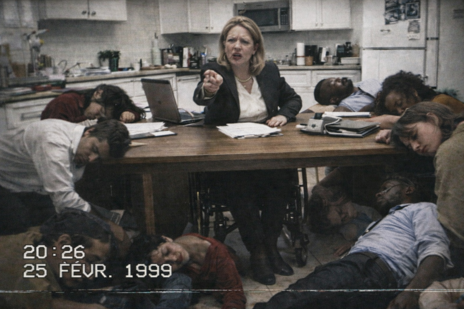 Une personne en fauteuil roulant, en tenue de bureau, est assise au centre d’une grande table de cuisine encombrée de documents et d’un ordinateur portable, pointant du doigt vers l’objectif. Autour de la table et au sol, plusieurs personnes paraissent inconscientes, affalées sur les chaises ou allongées, tandis qu’un encart de type enregistrement vidéo affiche l’heure « 20:26 » et la date « 25 févr. 1999 » en bas de l’image.