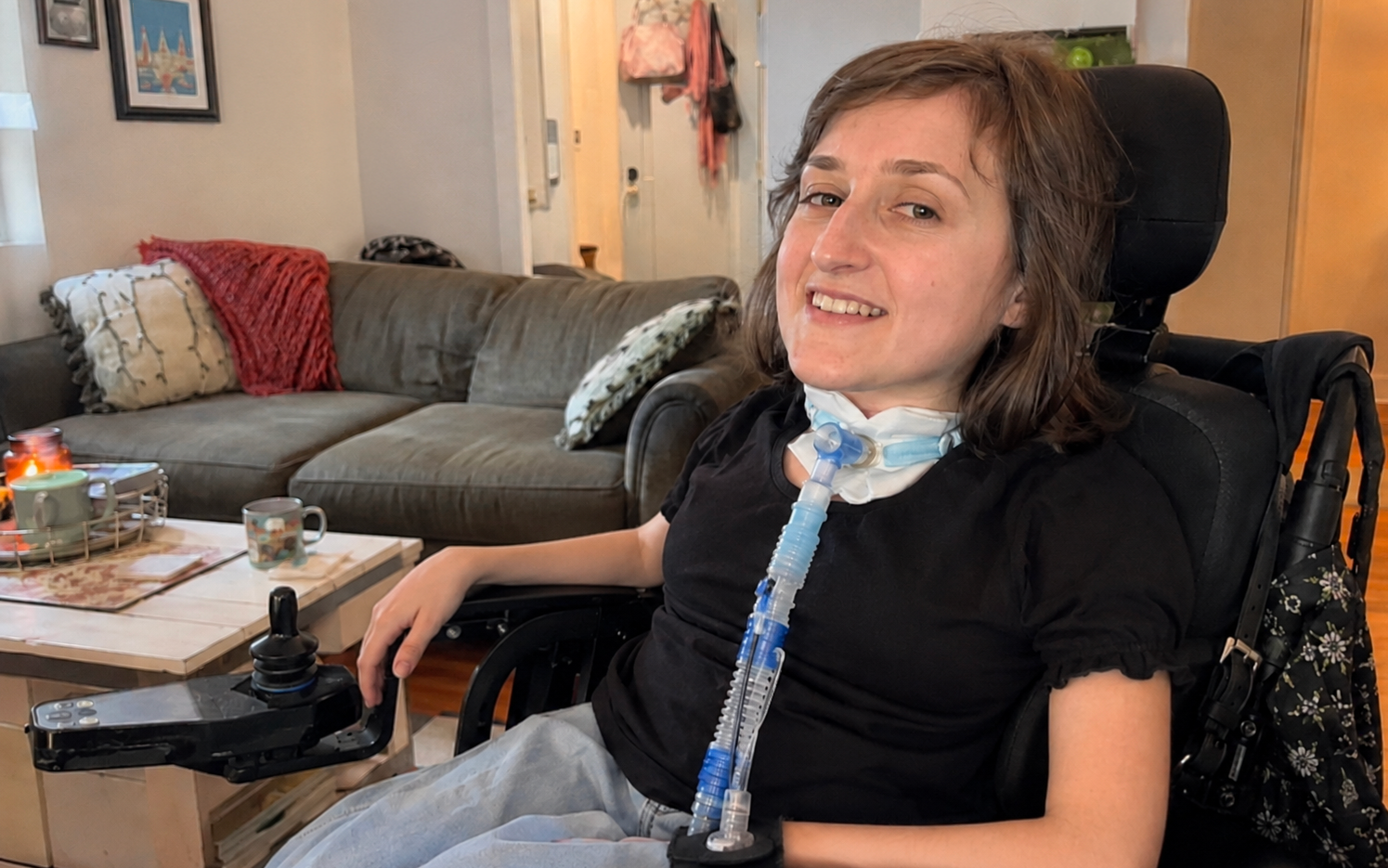 Portrait d'une femme souriante assise dans un fauteuil roulant électrique à l'intérieur d'un salon. Elle est équipée d'un tube respiratoire (trachéotomie) visible au niveau de son cou.