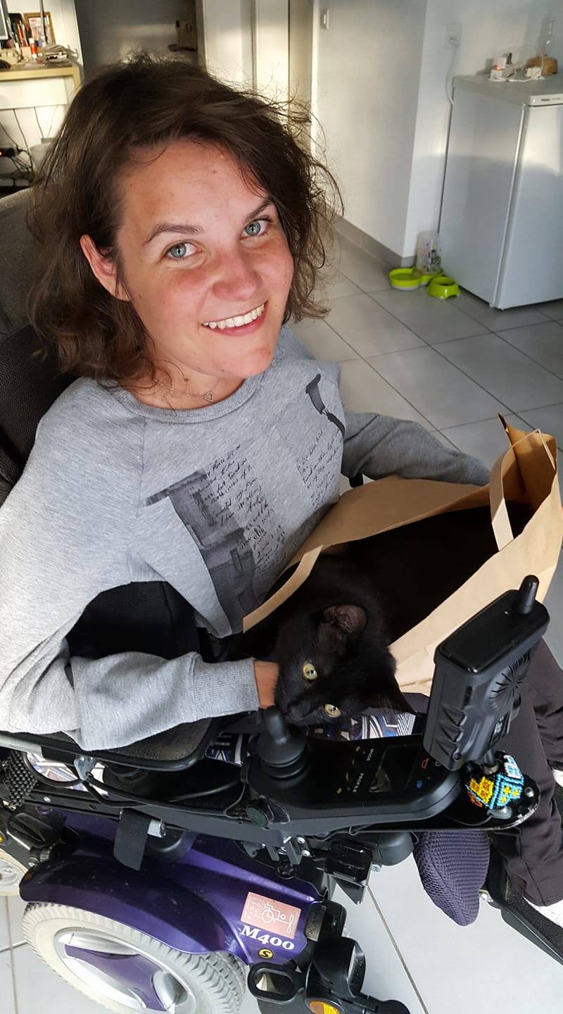 Pamela Ruga, en fauteuil roulant électrique, sourit, un chat noir glissé dans un sac en papier kraft sur ses genoux.