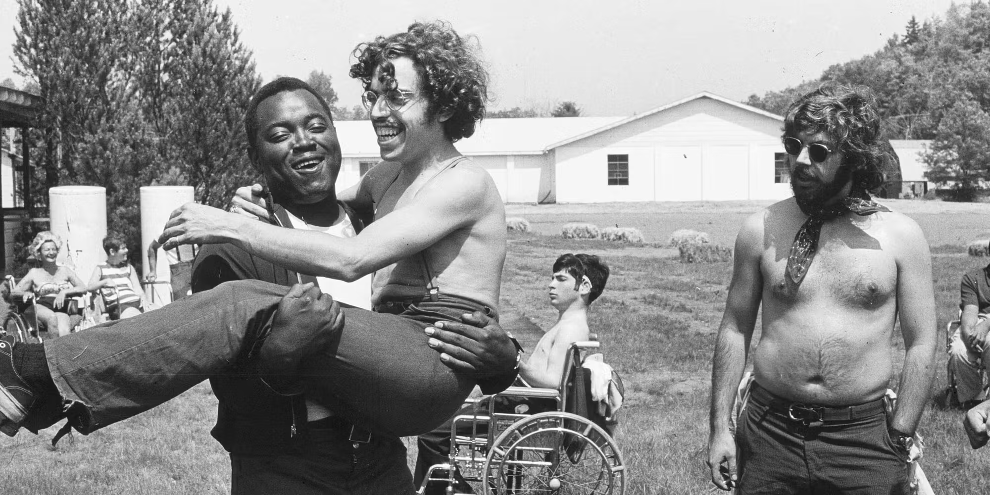 Photographie en noir et blanc, en plein air. Au premier plan, un jeune homme porte dans ses bras un autre jeune homme à lunettes rondes et cheveux bouclés, tous deux riant. En retrait sur la pelouse, devant de longs bâtiments bas, un jeune homme torse nu est assis dans un fauteuil roulant ; un homme barbu à lunettes de soleil et foulard se tient debout à droite.