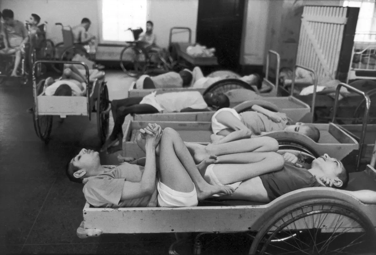 Dortoir de l'un des bâtiments de la Willowbrook State School. Une dizaine d'enfants et d'adolescents, torse nu ou en sous-vêtements, sont allongés sur des brancards à roues et des lits en bois montés sur roues de fauteuils roulants. Au premier plan, deux garçons reposent tête-bêche sur un même brancard, membres entremêlés. À l'arrière-plan, d'autres enfants sont assis dans des fauteuils roulants près d'une fenêtre. Photographie en noir et blanc.