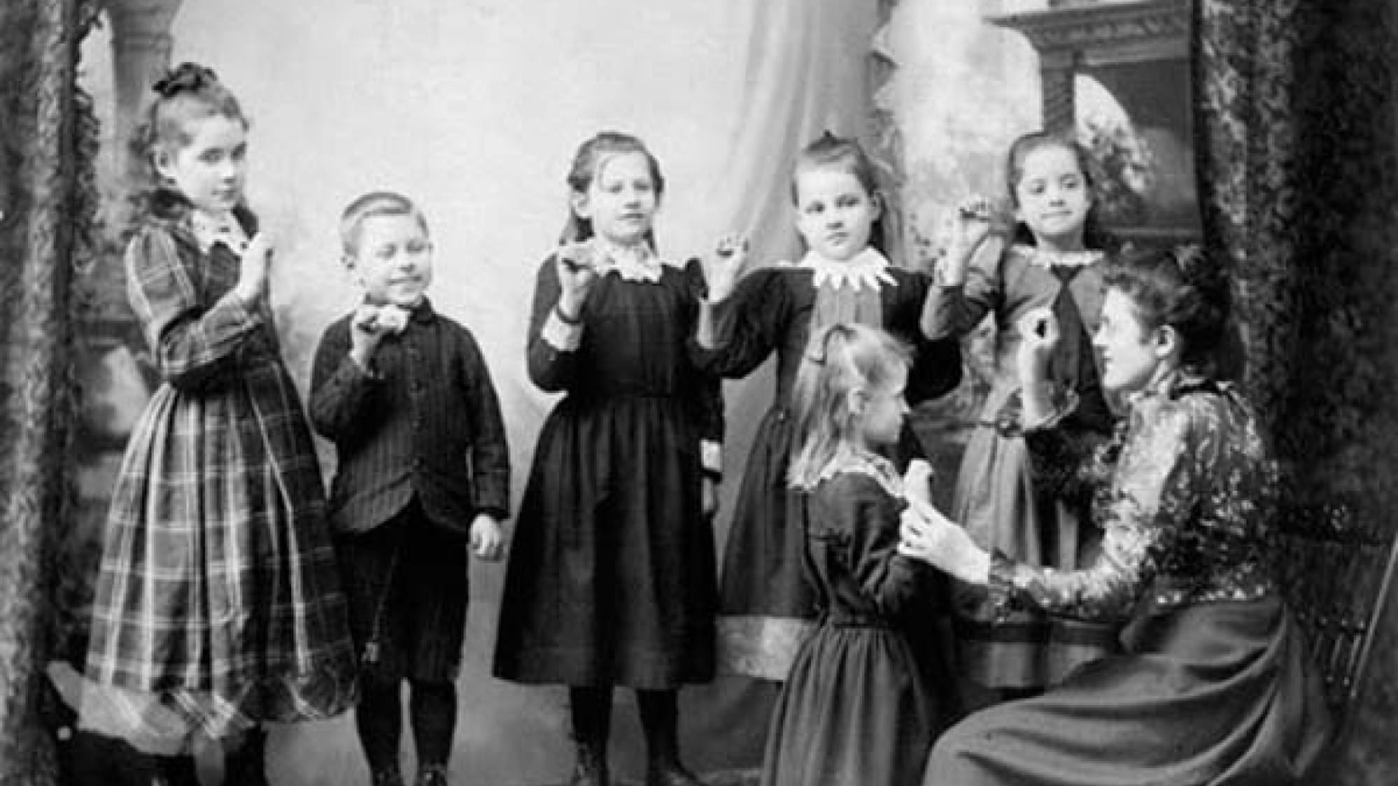 Photographie ancienne en noir et blanc. Une femme s'adresse en langue des signes à un groupe d'enfants, filles et garçons en vêtements de la fin du 19e siècle; plusieurs ont les mains levées, en train de signer à leur tour.