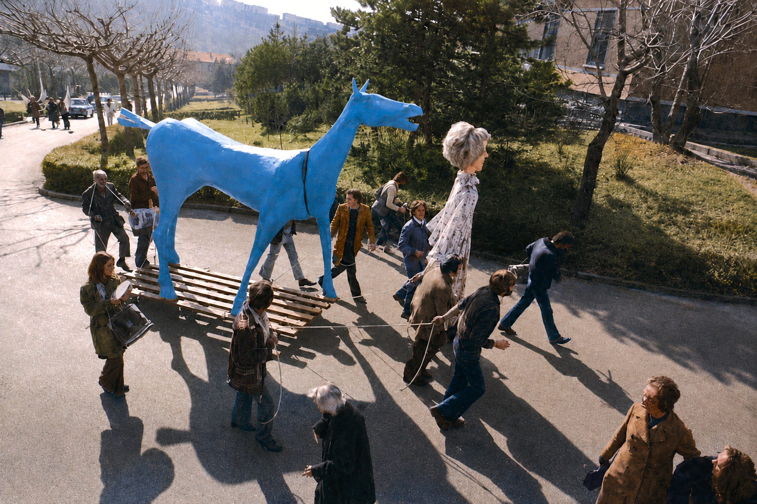 Vue en plongée d'une dizaine de personnes tirant avec des cordes une grande sculpture de cheval peinte en bleu vif, posée sur une plateforme de lattes de bois, le long d'une allée goudronnée bordée d'arbres. En manteaux et vestes, elles avancent vers la droite, une voiture claire et des bâtiments en arrière-plan.