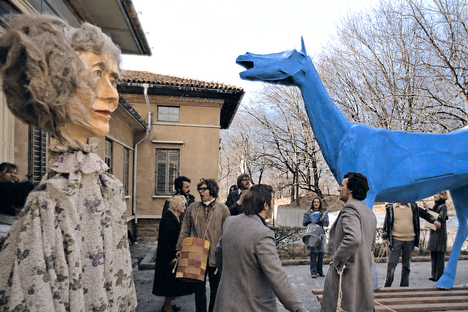 À gauche, une figure géante représentant le buste d'une femme âgée aux cheveux gris ébouriffés, vêtue d'une robe à motifs floraux ; à droite, la tête et l'encolure d'une grande sculpture de cheval peinte en bleu vif, gueule ouverte. Entre les deux, un groupe de personnes en manteaux et vestes, dont une tenant un tambour, devant un bâtiment aux volets et au toit de tuiles, sous des arbres dénudés.