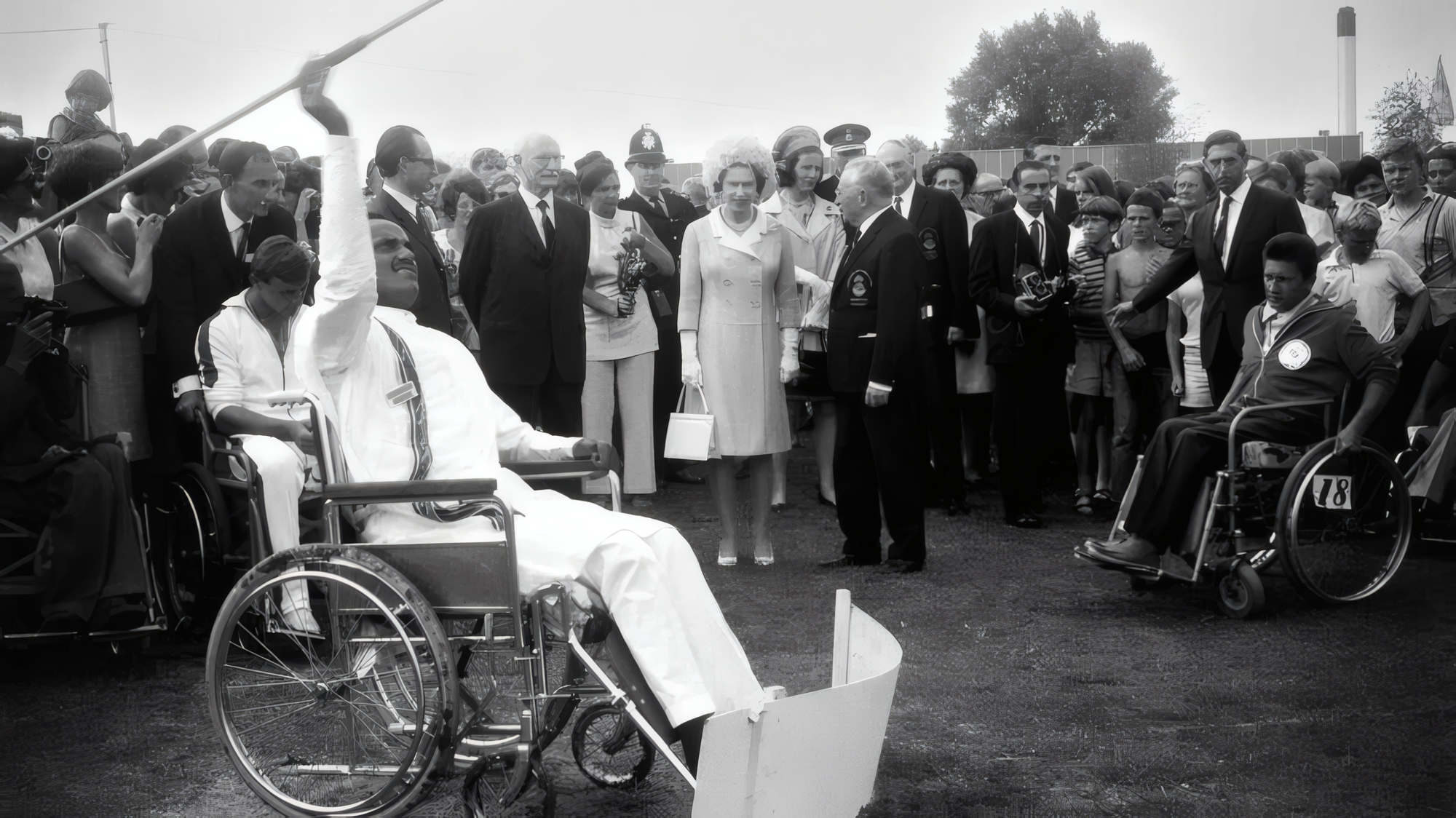 Photographie en noir et blanc. Sur une pelouse, un homme en tenue blanche assis dans un fauteuil roulant lève le bras et tient un javelot au-dessus de sa tête, prêt à lancer. La reine Élisabeth II, en tailleur clair et gants blancs, se tient parmi les spectateurs et le regarde. Autour d'elle, des hommes en costume sombre, un policier britannique en casque, d'autres femmes et des enfants. À droite, un homme en veste de survêtement, assis dans un fauteuil roulant, porte un dossard numéroté 18.