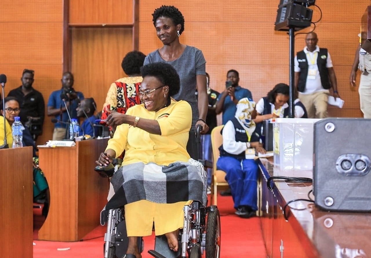Dans un hémicycle parlementaire aux murs de bois orangé, la députée Laura Kanushu, représentante nationale des personnes en situation de handicap, se déplace en fauteuil roulant manuel, poussée par une assistante en robe grise. Vêtue d'une robe jaune, elle sourit et lève la main droite en signe de salut. Autour d'elle, des huissiers en uniforme, des journalistes et du personnel de sécurité observent la scène près des bancs et pupitres de la salle.
