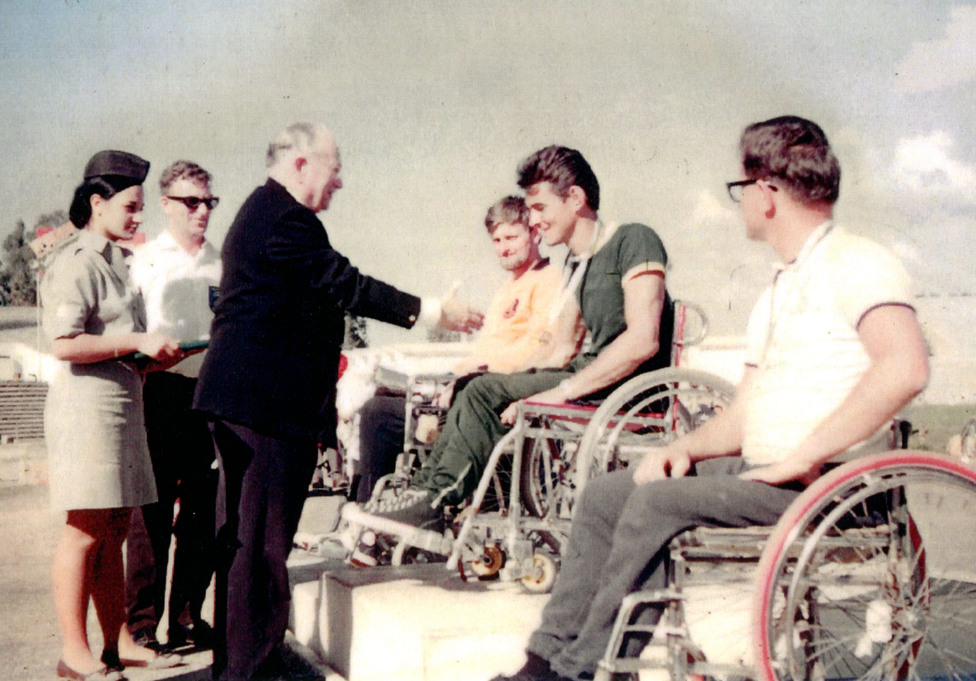 Photographie couleur ancienne. En extérieur, un homme âgé en costume sombre (Ludwig Guttmann) tend la main vers deux jeunes hommes assis dans des fauteuils roulants à cadre chromé, l'un en maillot orange, l'autre en maillot vert, une médaille au cou. À l'arrière-plan, une femme en uniforme clair et coiffe tient un plateau, à côté de deux hommes debout. À droite, un autre homme en fauteuil roulant, en polo blanc et lunettes, observe la scène.