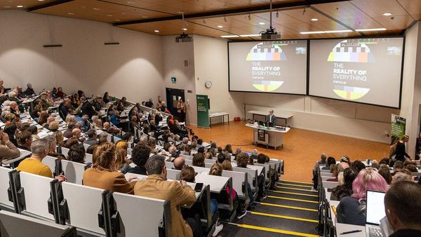 Crowded lecture theatre at the Reality of Everything symposium. MC Professor Huhana Smith is standing behind the lectern at the front.