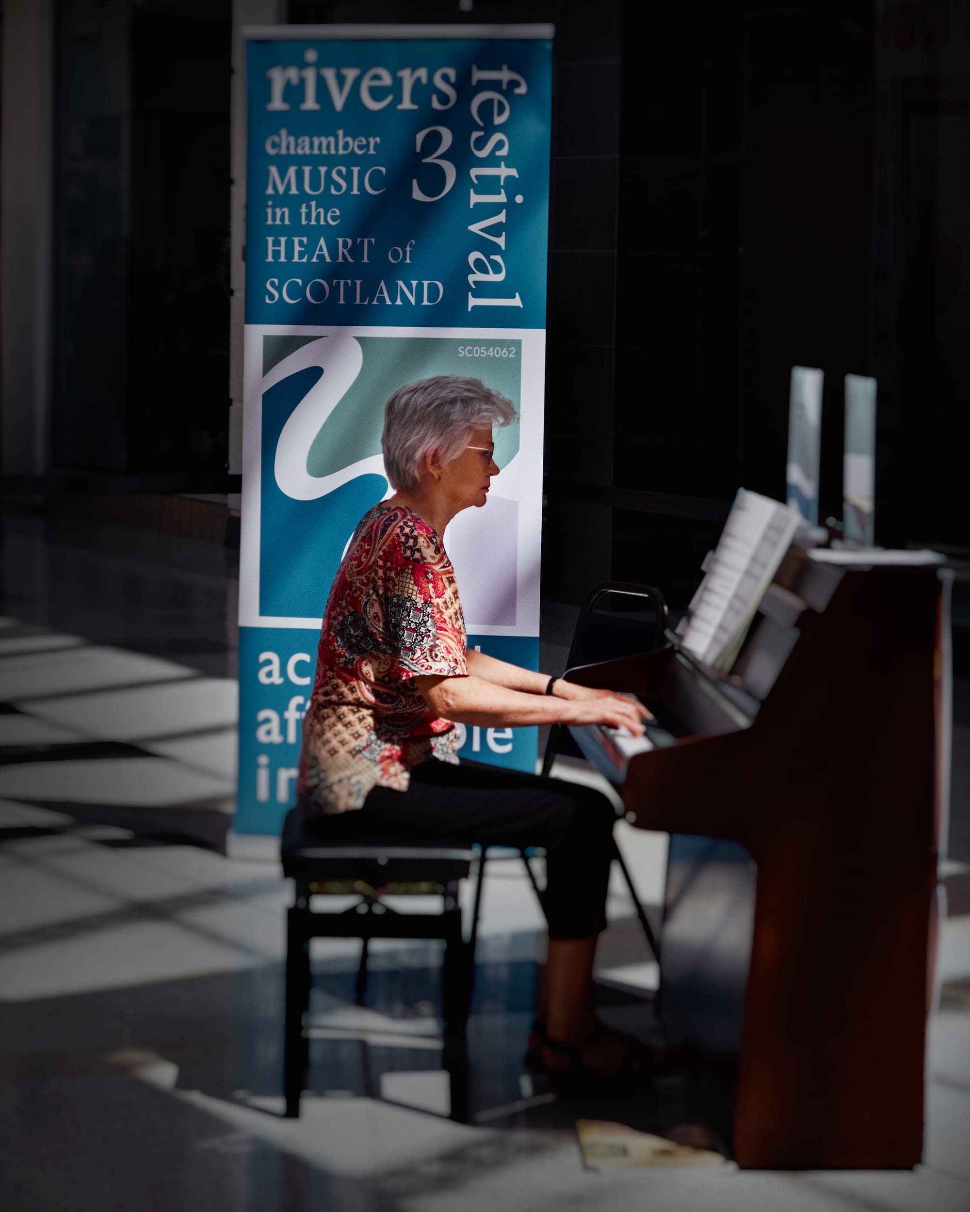 Kirsty playing piano in dappled light in front of a large banner advertising the Three Rivers music festival.