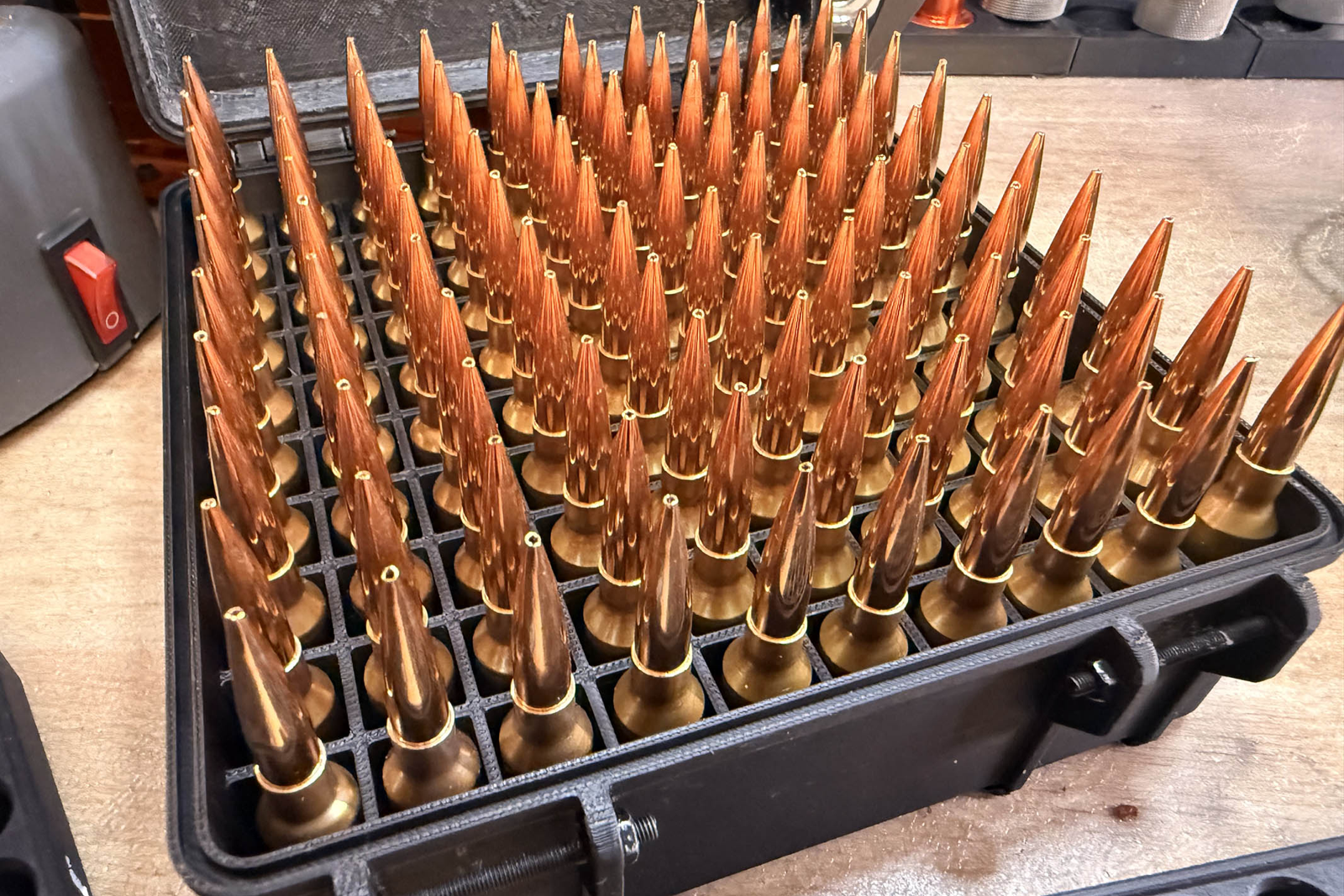 A black ammunition box holding rows of completed handloaded cartridges on a work surface.