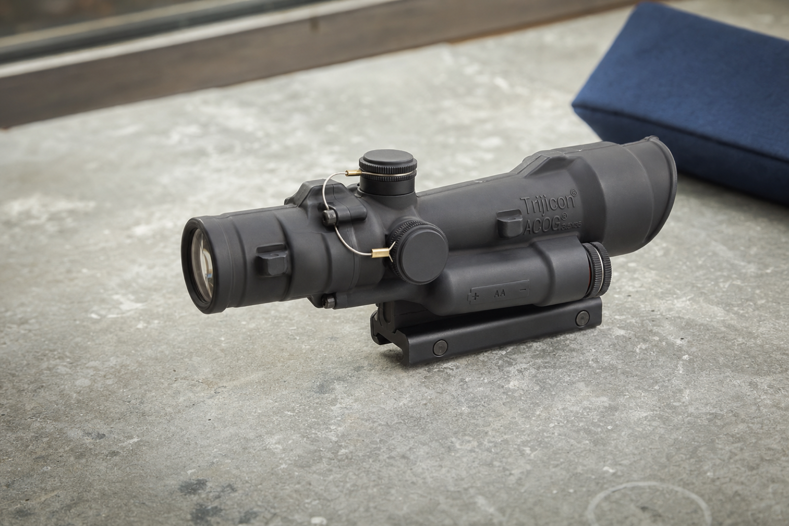 A black LED ACOG rests on its mounting foot, with its battery housing and capped controls visible from the side.