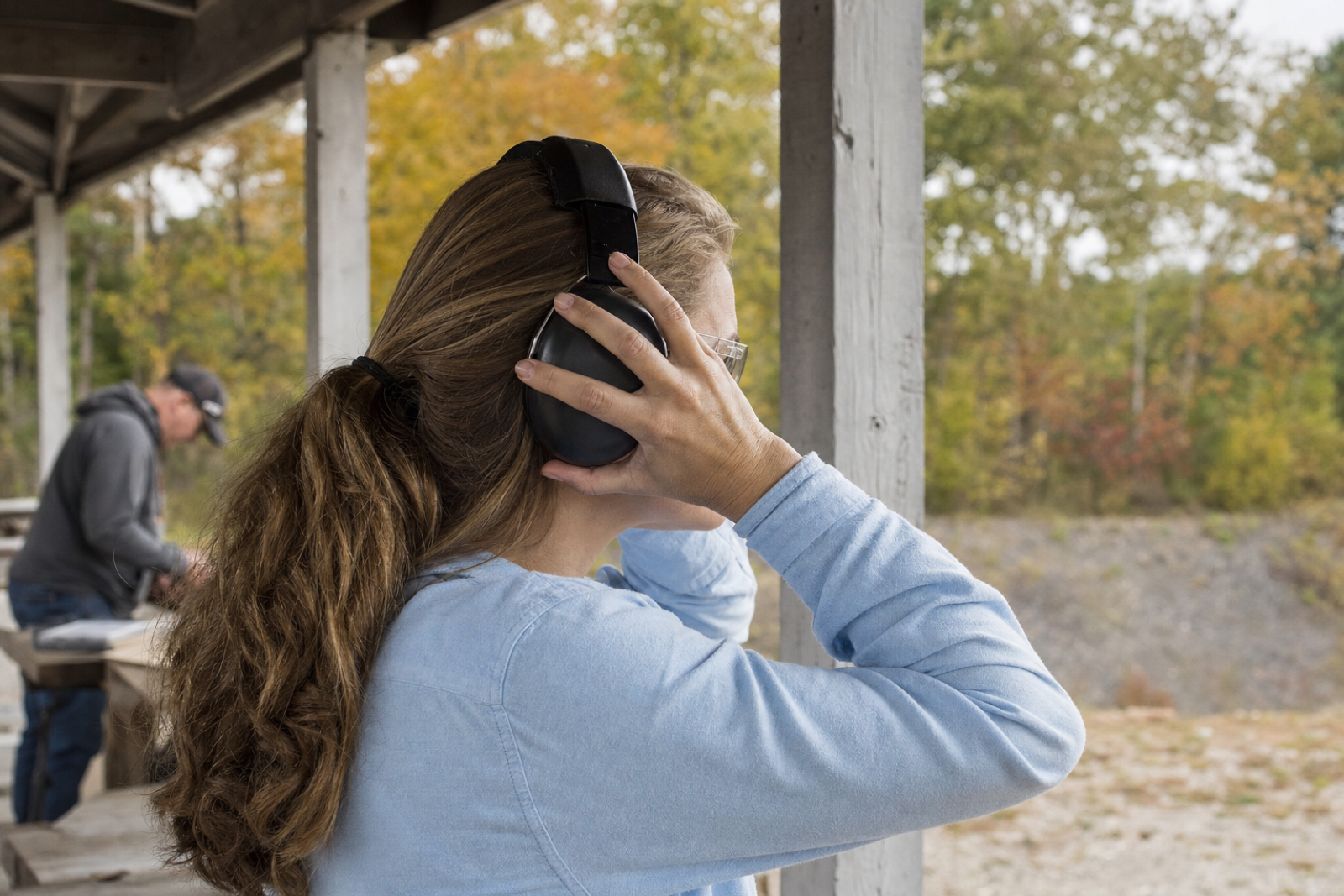 A club participant in a pale blue shirt adjusts dark hearing protectors beside a timber range shelter.