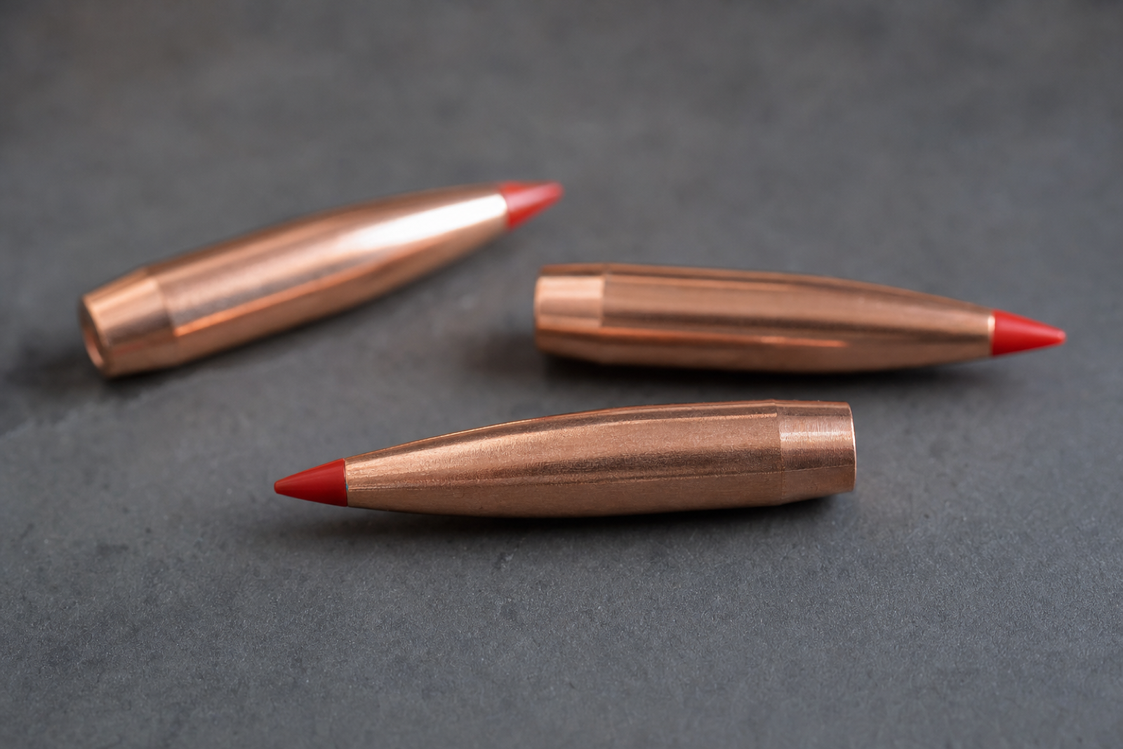 Three loose copper-jacketed bullets with red polymer tips on a dark grey surface.