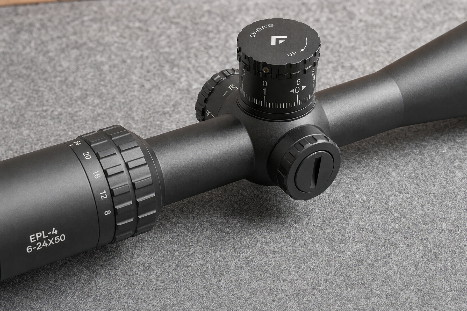 Close view of a black scope’s elevation turret, slotted side control and connected tube on a grey mat.