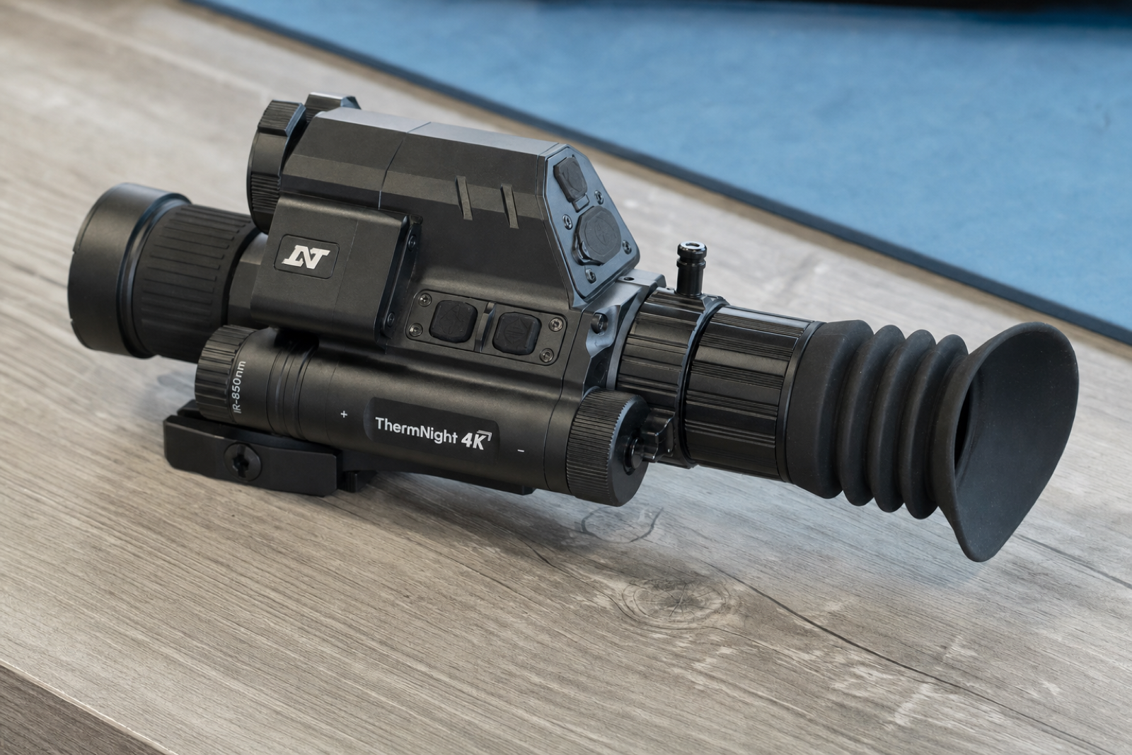 A side and rear view of the DNT ThermNight TNC4K219R shows its rubber eyecup, ribbed adjustment ring, and connected control housing on a grey wooden work surface.