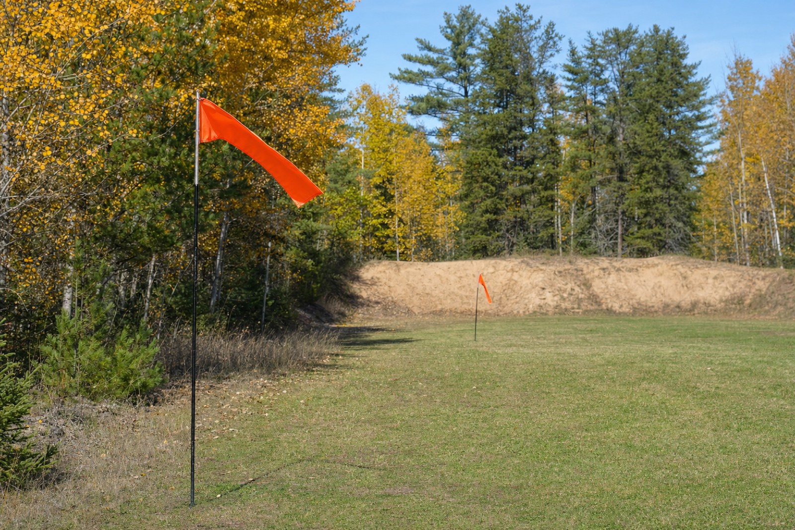 Two orange wind flags stand at different distances beside a grass range lane and an autumn tree line.