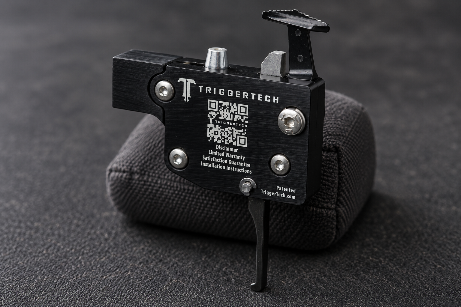 The reverse side of a TriggerTech Tikka Special trigger assembly rests on a padded support, with the housing and flat lever visible.