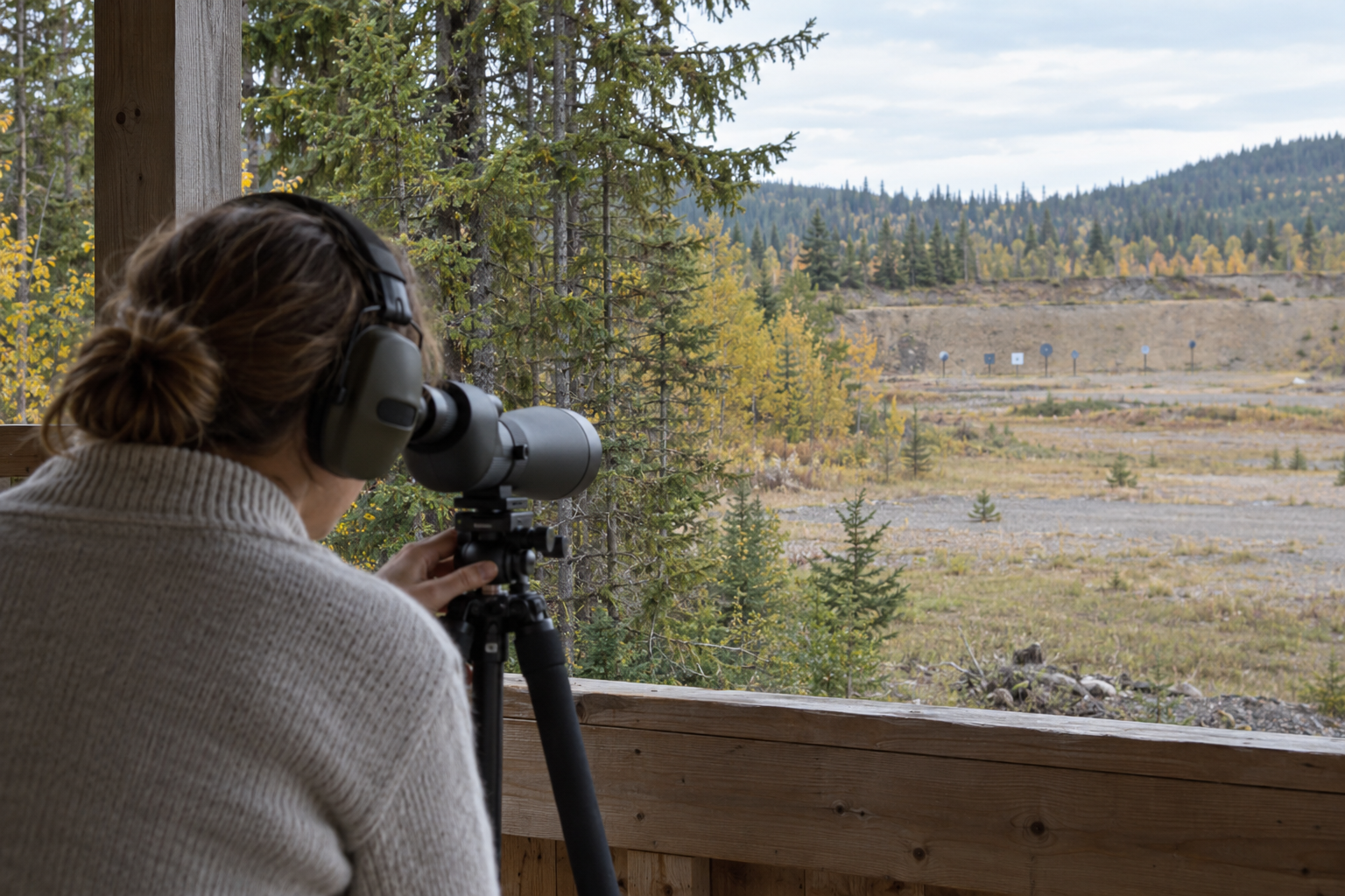 A person wearing earmuffs looks through a tripod-mounted spotting scope toward steel targets.