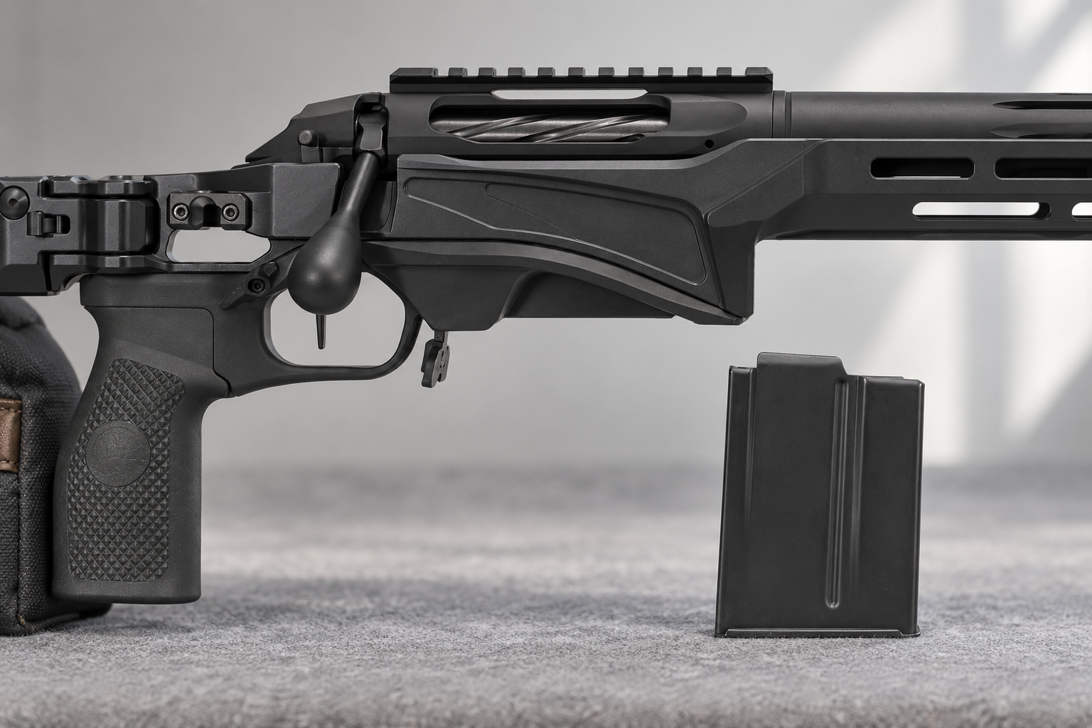 A detached rifle magazine stands on a bench mat beside the magwell of a supported chassis rifle.