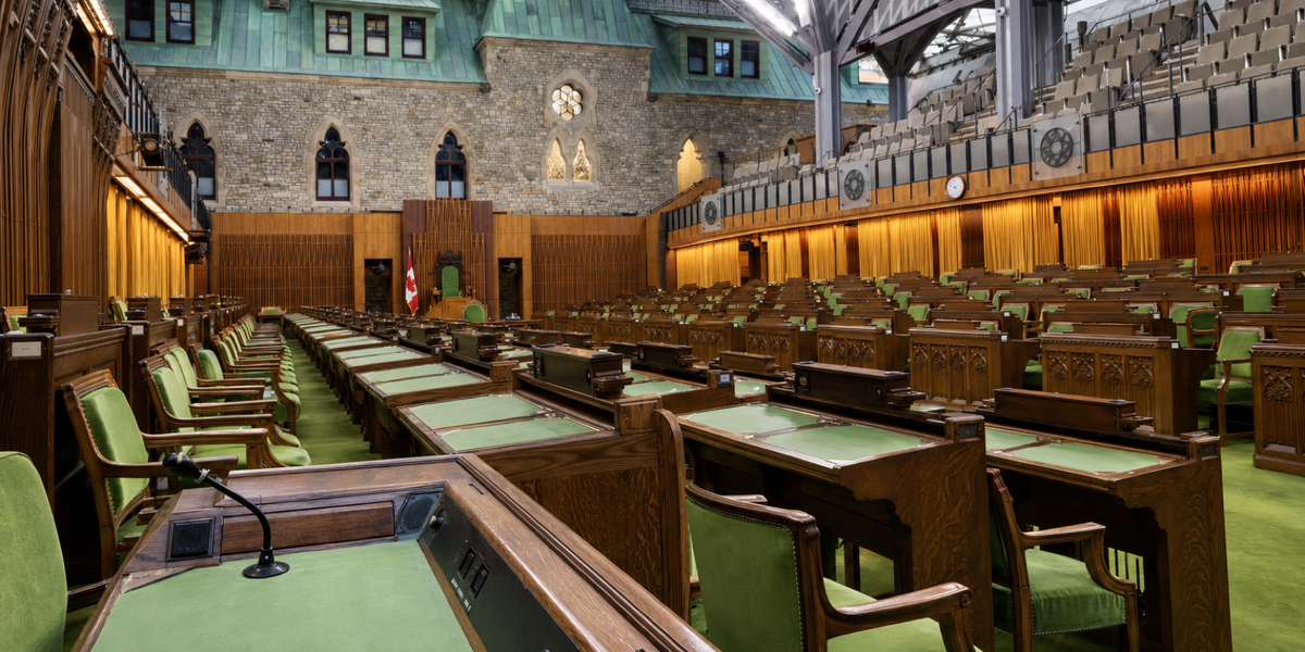 An empty Commons chamber with green seats and oak desks facing across a central aisle beneath a modern glass roof.