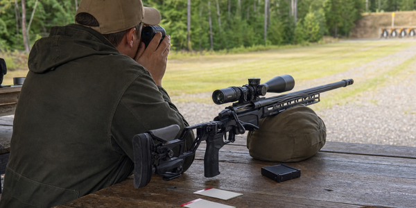 A shooter ranges a target beside a supported rifle.