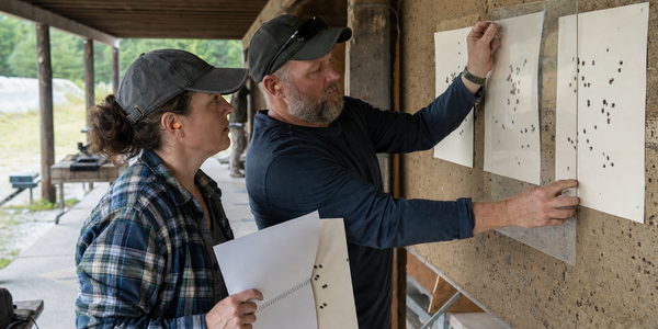 Two people compare several paper targets after a ceasefire at an outdoor range.
