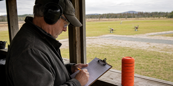 A range volunteer records a round on a clipboard beside a stack of orange clay targets.
