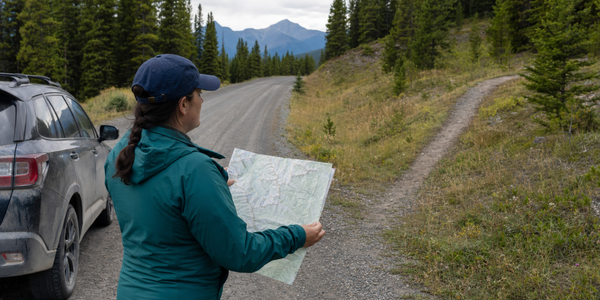 A public-land visitor compares a folded map with a gravel road and narrower recreation trail in the Alberta foothills.