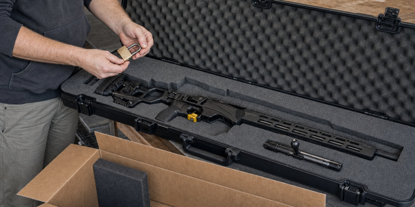 An unloaded bolt-action rifle with its bolt removed and trigger lock attached rests in an open hard case beside an anonymous shipping box.
