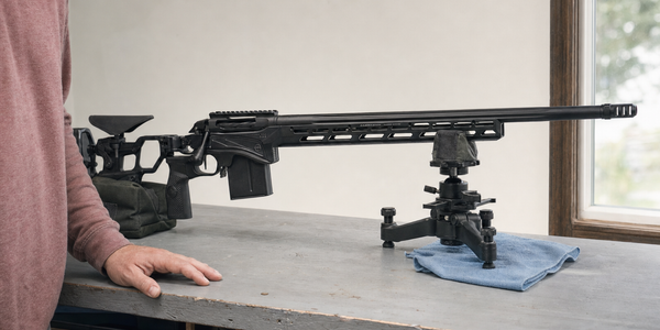 A supported modern chassis bolt-action rifle on a home workbench beside a partially visible owner.