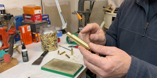 Two hands holding and examining an empty brass case above a workbench, with a jar of cases nearby.