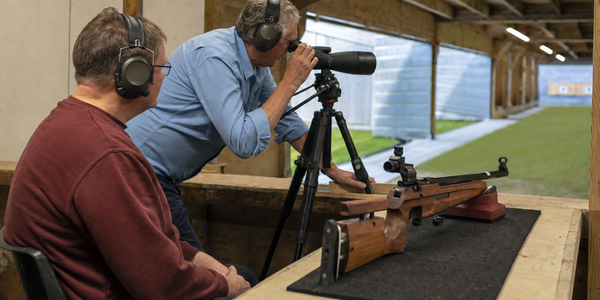 Generated illustration of a participant and volunteer at an indoor smallbore firing point with a supported target rifle.