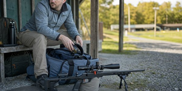 A seated competitor pauses with hearing protection partly out of a range bag beside a supported scoped rimfire.