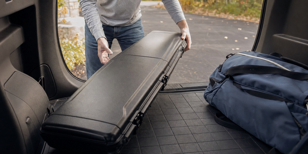 Hands guide a closed long protective equipment case into a vehicle cargo bay beside an overnight bag.