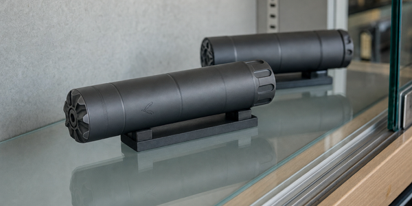 Two dark cylindrical suppressors rest on a pale shelf inside a glass display.