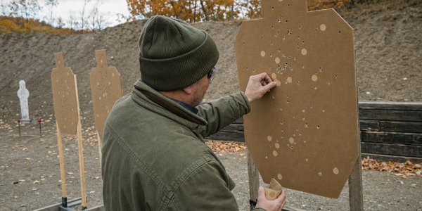 A volunteer patches holes in a brown cardboard target with tan pasters on an outdoor shooting bay, with two more cardboard targets, a white steel target and autumn trees behind.