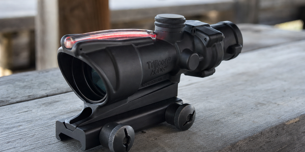 Close side view of a compact black ACOG optic with a red fibre-optic collector and mounting foot on a grey timber bench.